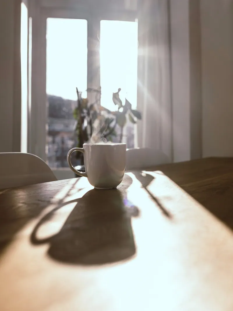 Caneca branca fumegante sobre uma mesa de madeira diante de uma janela ensolarada.