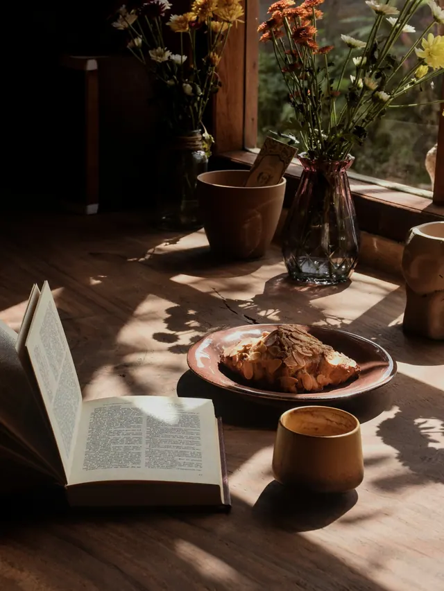 Livro aberto, croissant e xícara de café sobre uma mesa iluminada pelo sol.